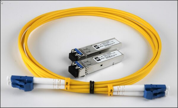 A yellow fiber optic cable with blue connectors and two silver SFP transceivers placed together on a white surface.